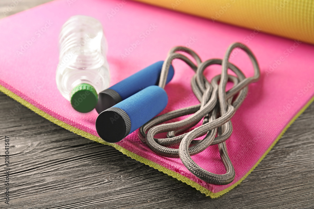 Yoga mat with jump rope and bottle of water on wooden background, closeup