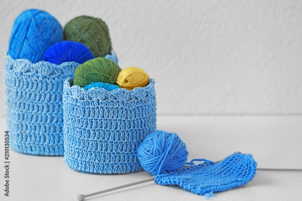 Basket with knitting yarn and unfinished clothes on table against light background