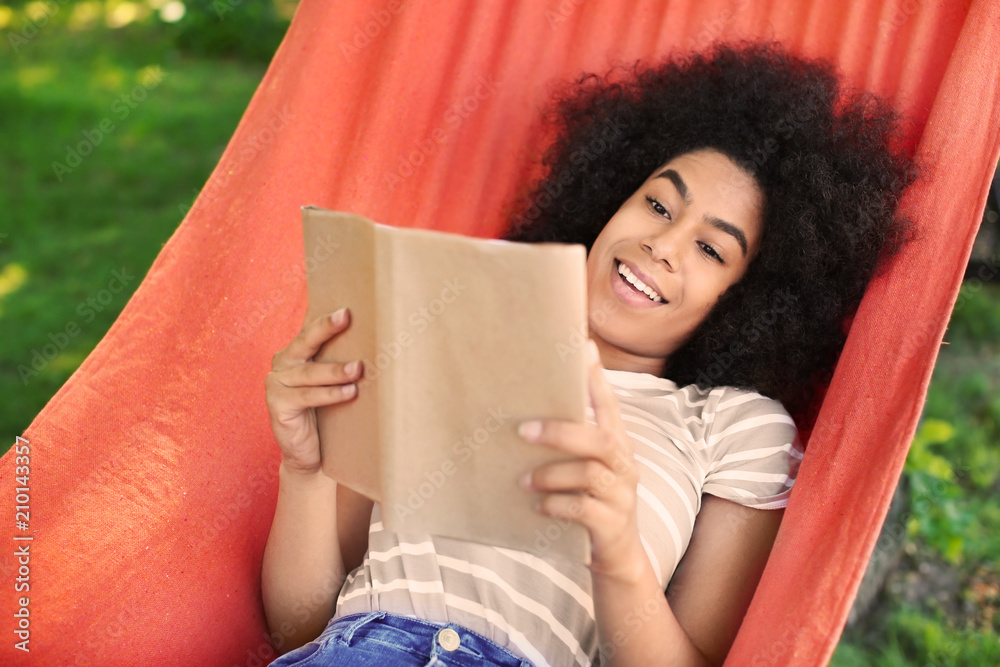 Beautiful young African-American woman reading book in hammock outdoors