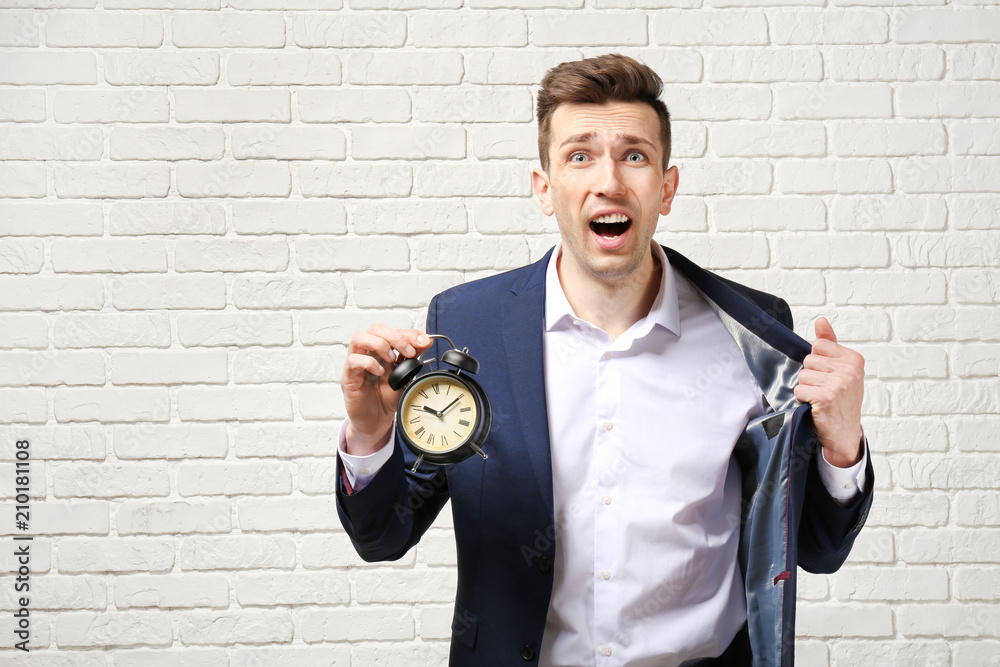 Troubled businessman with alarm clock against white brick wall. Time management concept