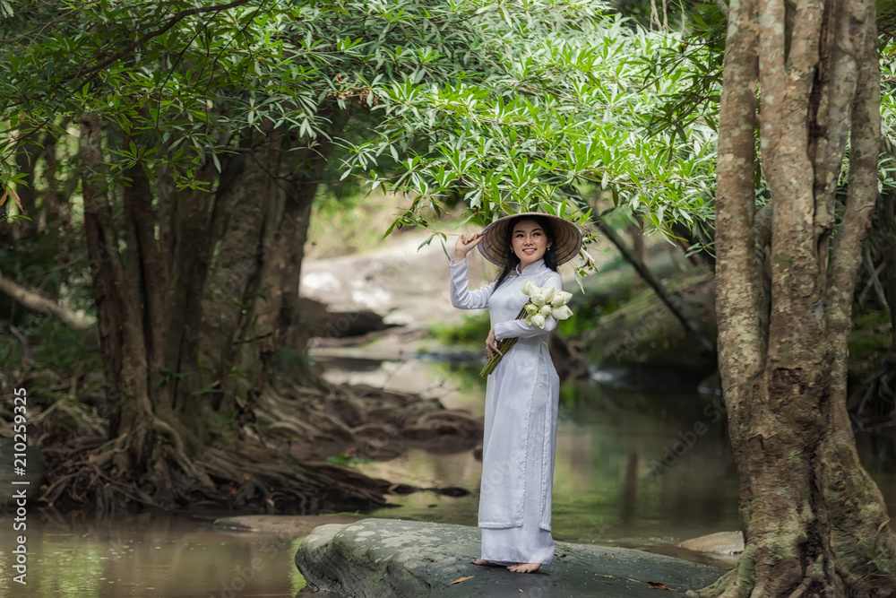 美丽的女人戴着帽子穿着敖黛传统的越南连衣裙站在森林里。