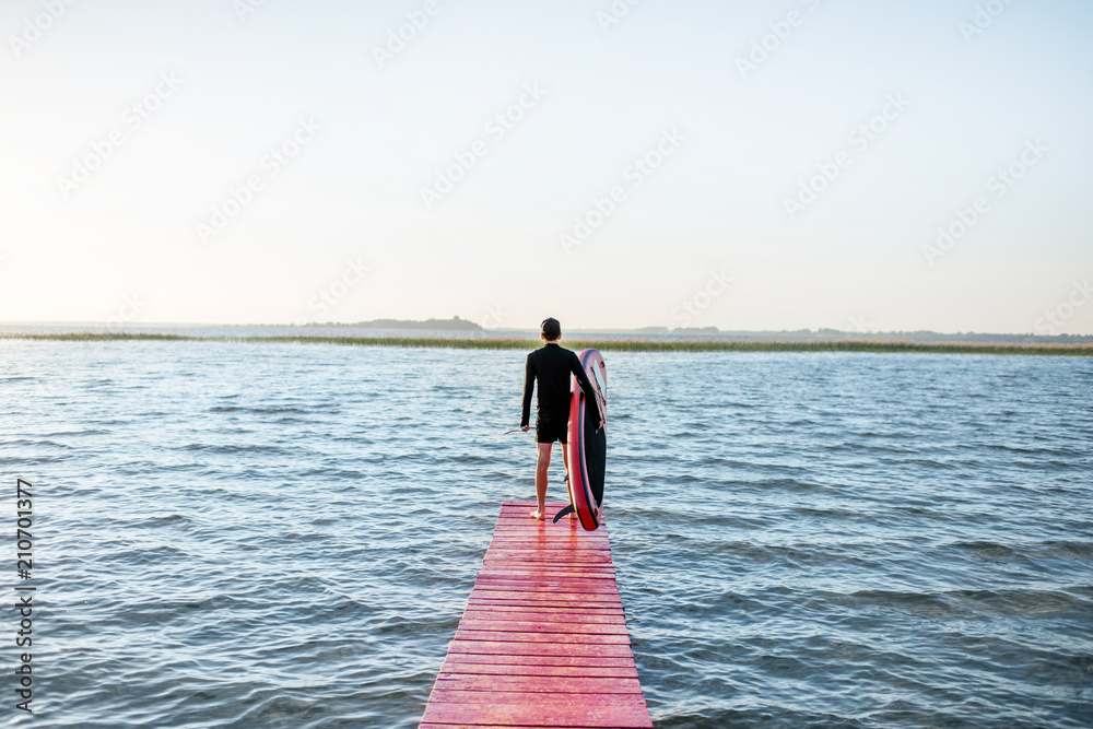 Landscape view on the lake with man standing with paddleboard on the pier during the sunrise