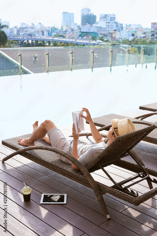 Young asian man reading book by the pool on a sunny summer day