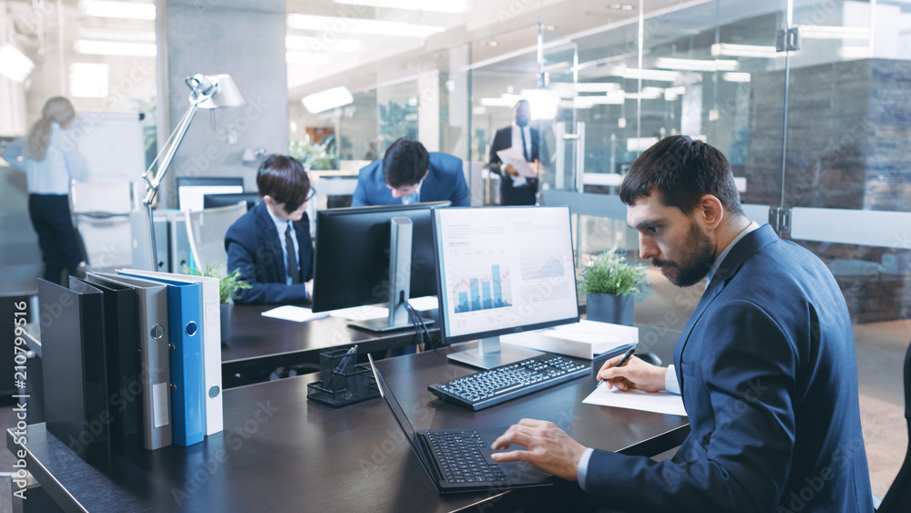 Professional Businessman Works on His Desktop Computer, Uses Laptop. In the Background Busy Office w