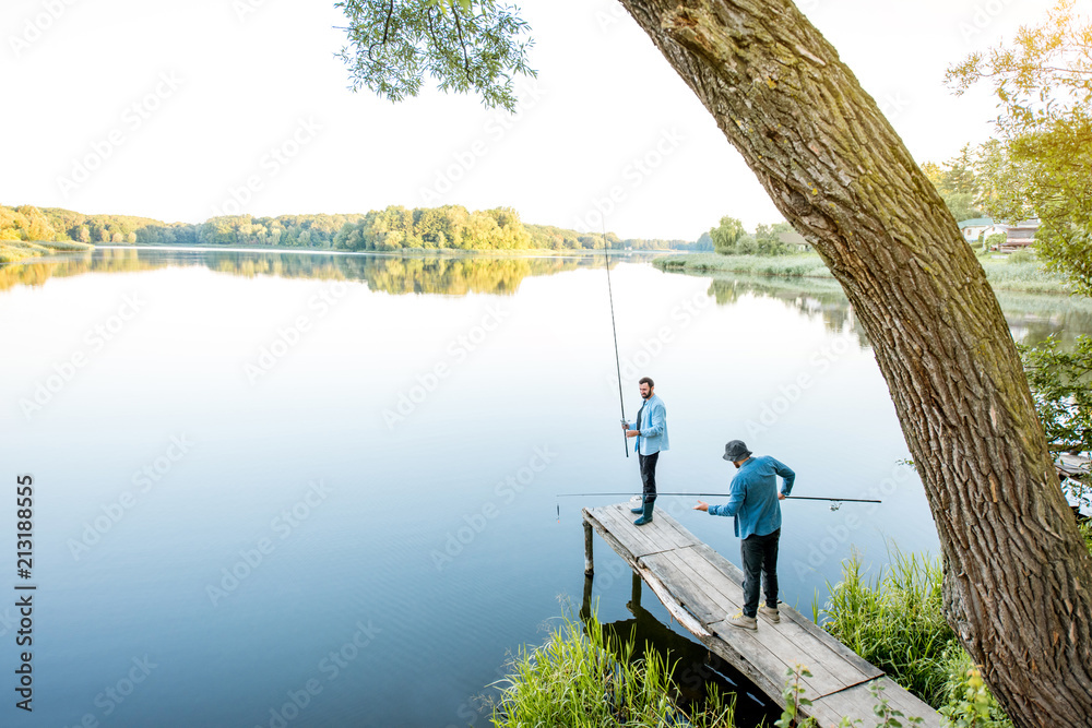 Landscape view on the beautiful lake with two male friends fishing together standing on the wooden p