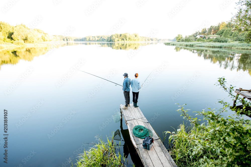 Landscape view on the beautiful lake with two male friends fishing together standing on the wooden p