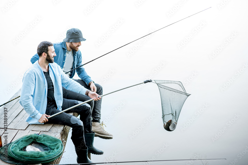 Two friends catching fish with fishing net and rod sitting on the wooden pier at the lake