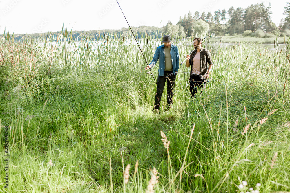 Two fishermen walking with fishing rod and net on the green lawn near the lake in the morning