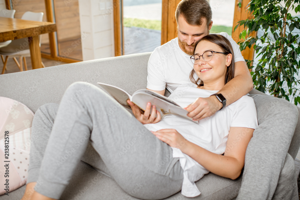 Young lovely couple dressed in pajamas reading books hugging together on the couch in their cozy hou