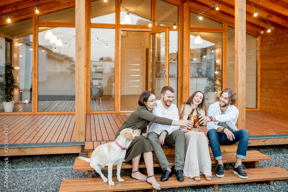 Young friends enjoying evening time sitting together with dog on the terrace of the modern wooden ho