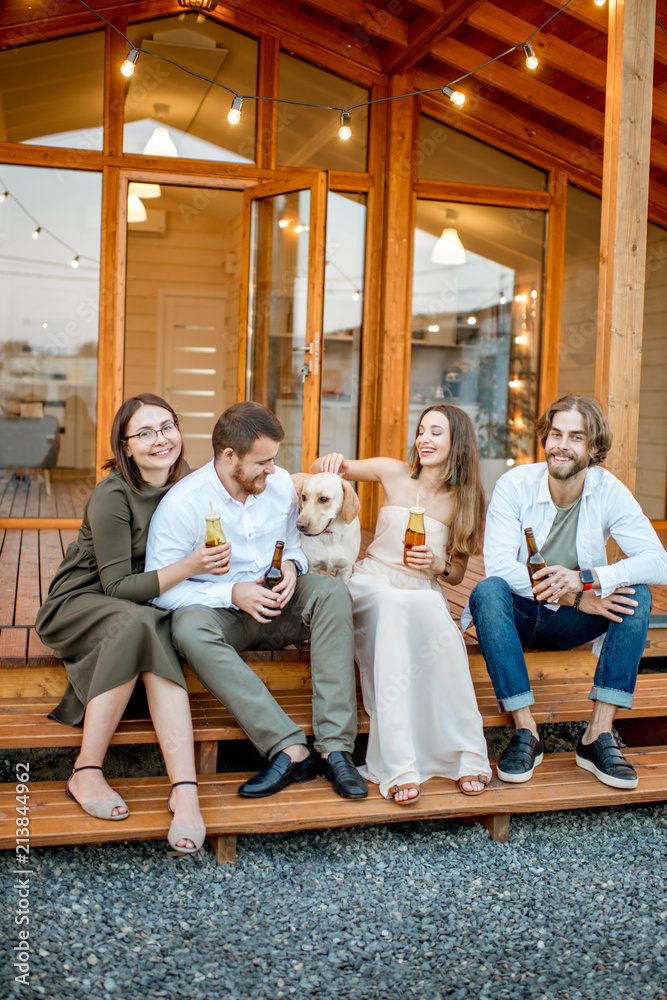 Young friends enjoying evening time sitting together with dog on the terrace of the modern wooden ho