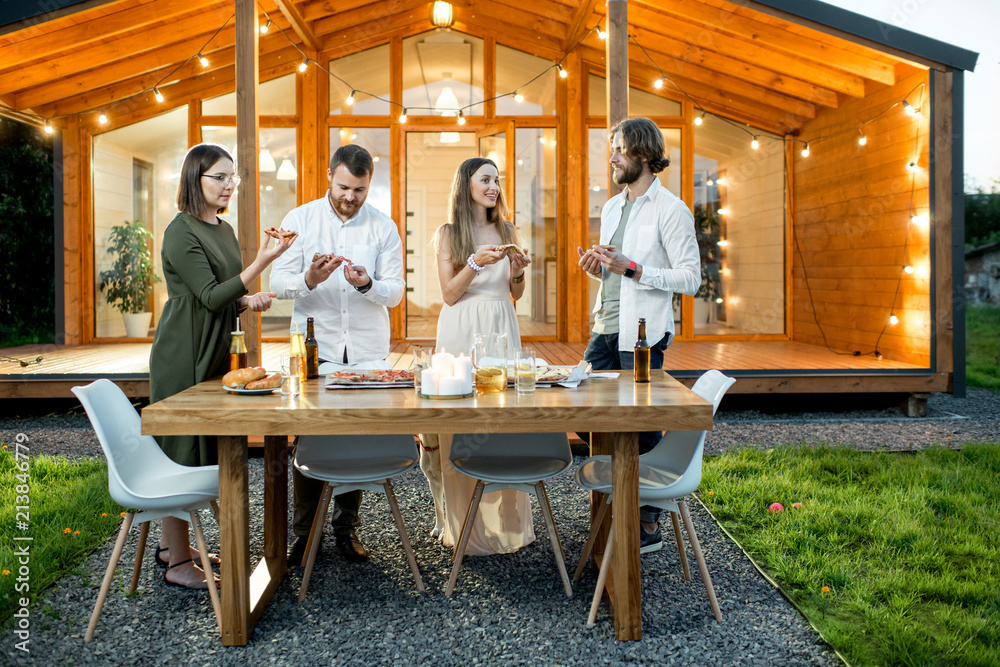 Friends having a dinner with pizza on the backyard of the modern house decorated with lights during 