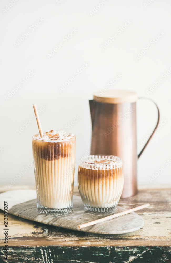 Iced coffee in tall glasses with milk and straws on board over rustic wooden table, white wall and j