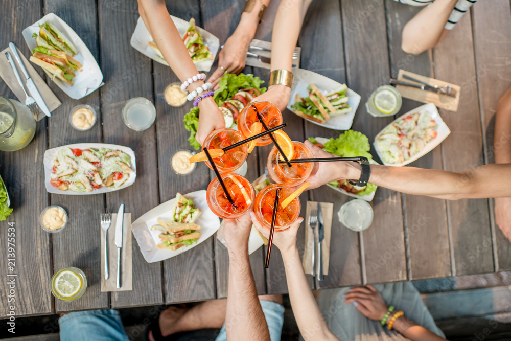 Friends clinking glasses with cocktail drinks, top view on the table full of snacks outdoors