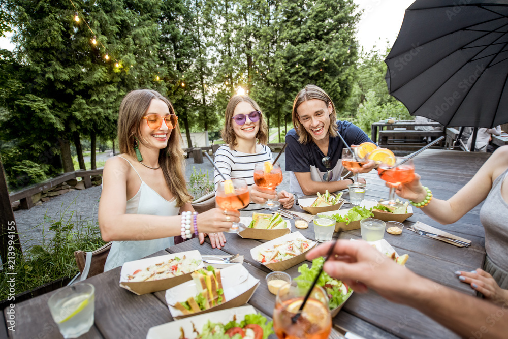 Young friends having fun sitting together with tasty snacks and drinks during the evening lights at 