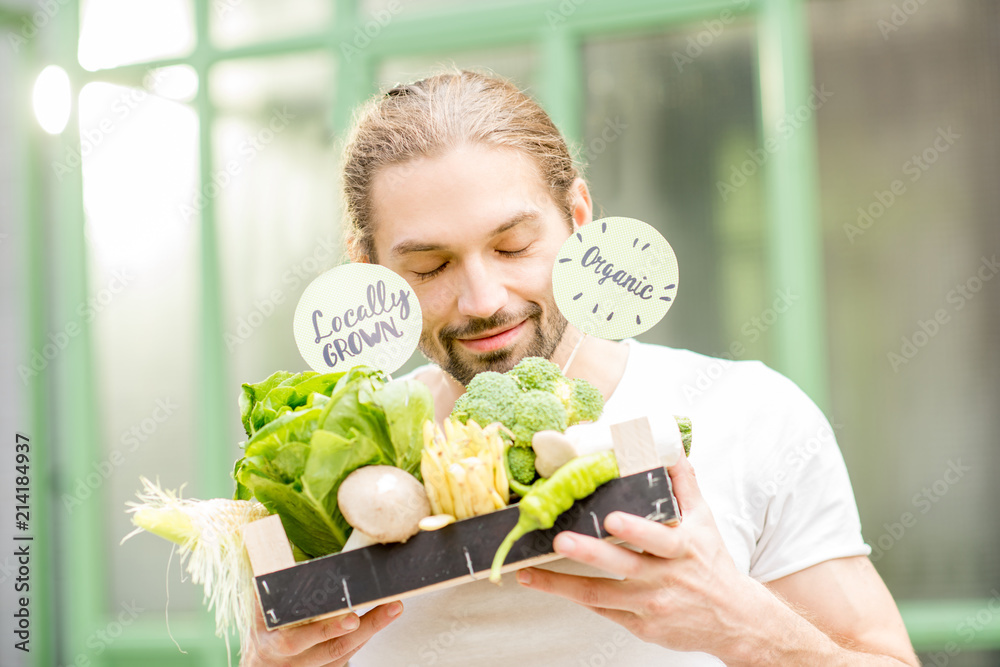 Portrait of a happy vegetarian man holding box full of fresh raw vegetables outdoors on the green ba