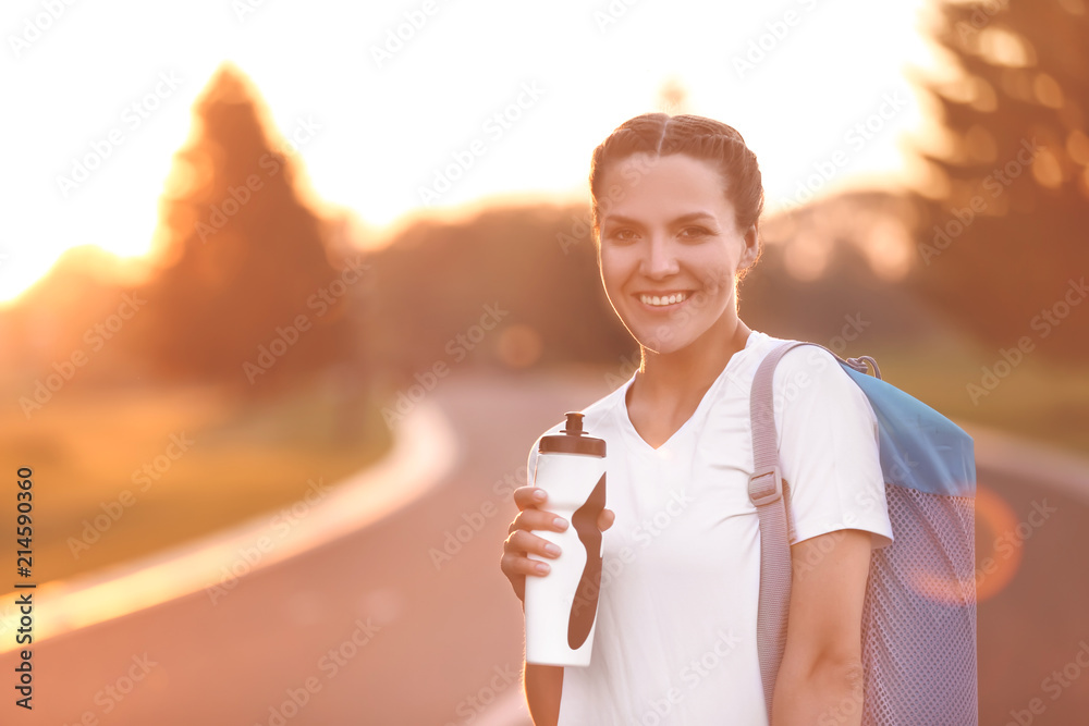 美丽的年轻女子，早上在户外拿着一瓶水和瑜伽垫