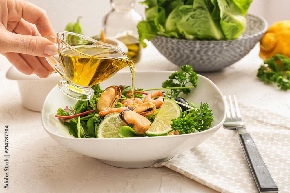 Woman pouring oil into bowl with fresh salad on table