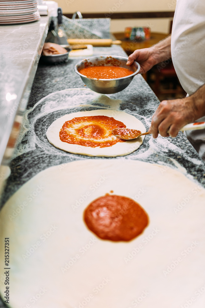 Raw pizza dough with tomato sauce. Chef adding sauce.