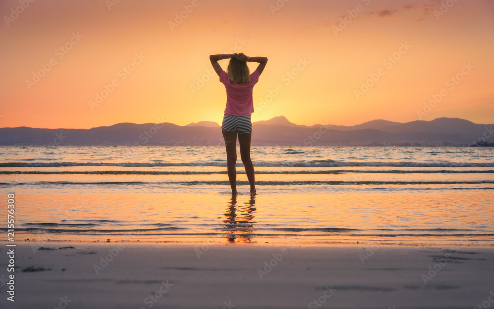 年轻的女人站在海边，沙滩上的海浪映衬着群山，夕阳下的橙色天空