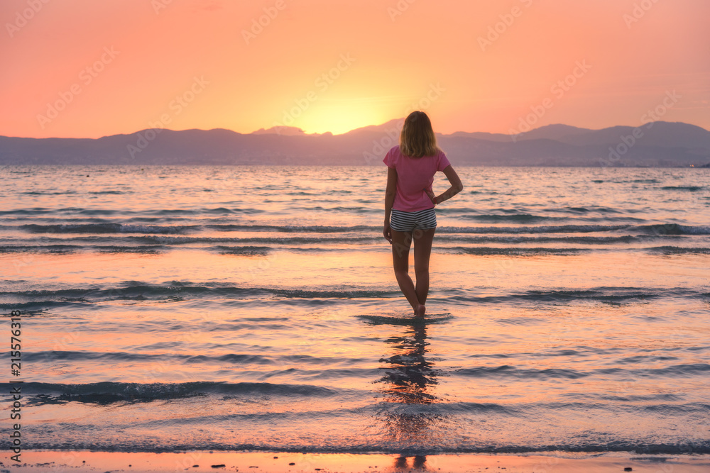 年轻女子站在大海中，沙滩上的海浪映衬着群山和日落时橙色的天空