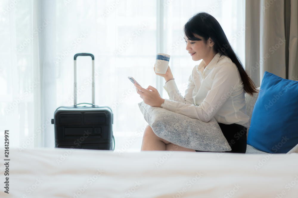 Beautiful Asian young smiling businesswoman in suit drinking coffee and using mobile phone while sit