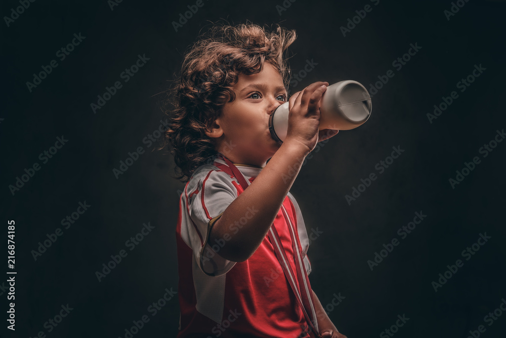 Little champion boy in sportswear with a gold medal drinking water from a bottle. Isolated on a dark