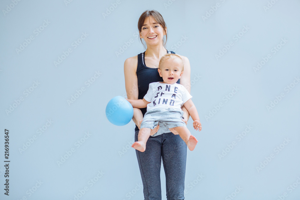 快乐的大笑妈妈在家里用灰色背景为蹒跚学步的男婴做体操。健身，哈