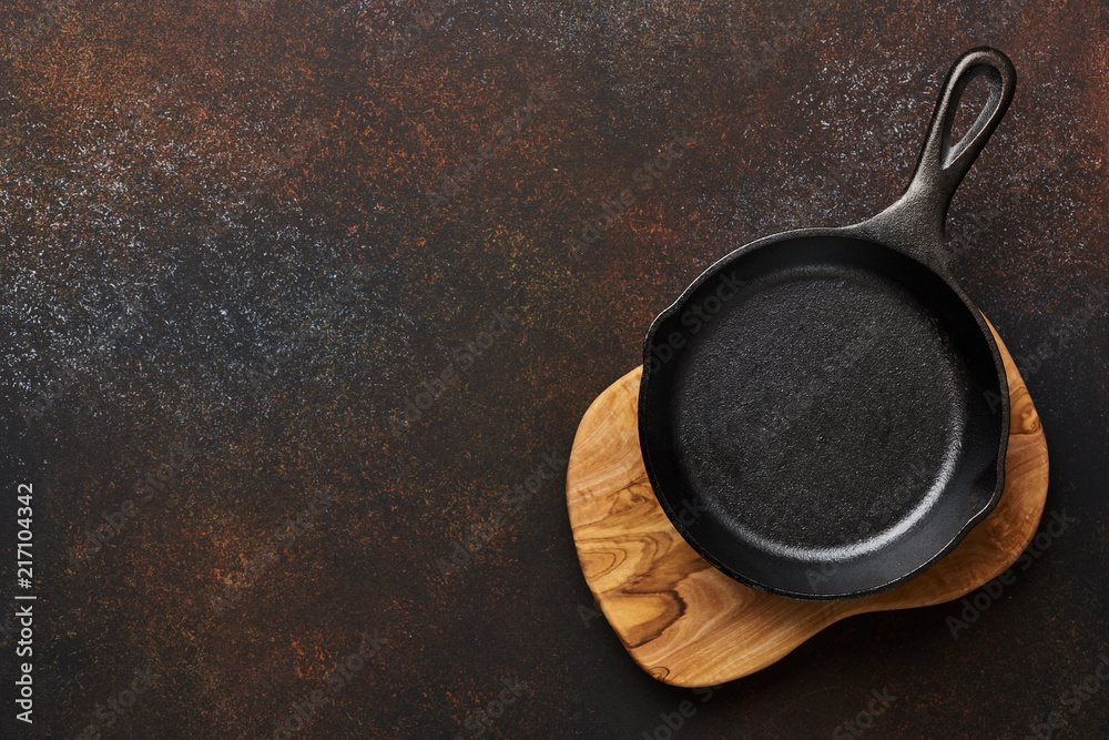 Blank black fry pan on olive wooden board over brown concrete background with copy space.