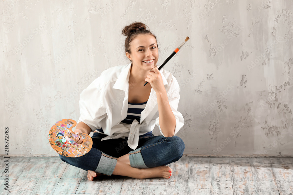 Female artist with brush and paint palette sitting on floor near grey wall