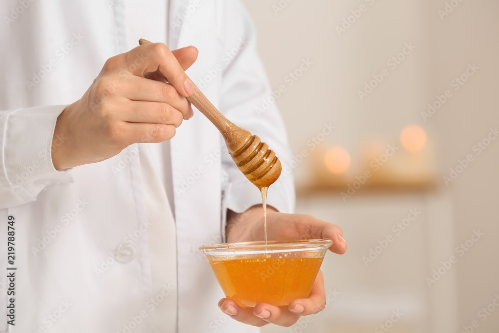 Female cosmetologist holding dipper and bowl with honey on blurred background