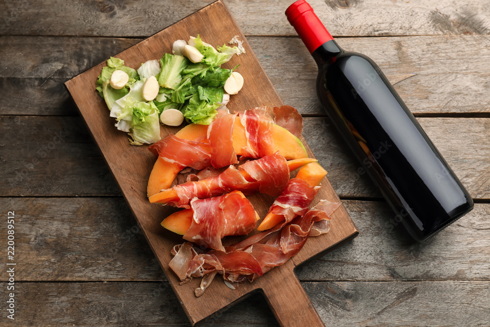 Board with delicious melon wrapped in prosciutto and bottle of wine on wooden table