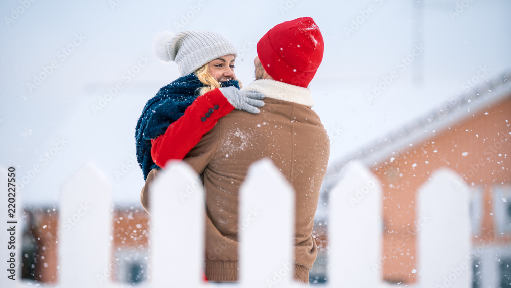 英俊的年轻情侣拥抱并幸福地微笑，雪落在他们身上。情侣在后院相爱