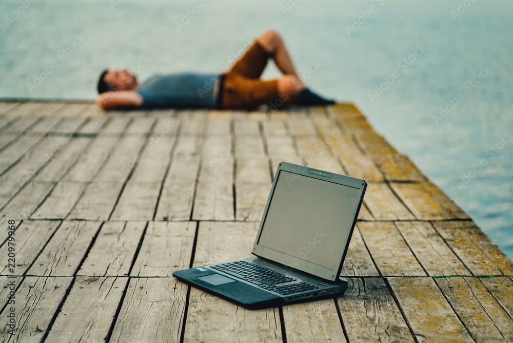 A young man lays on a wooden masonry near the river. He puts the working laptop away and enjoy by hi