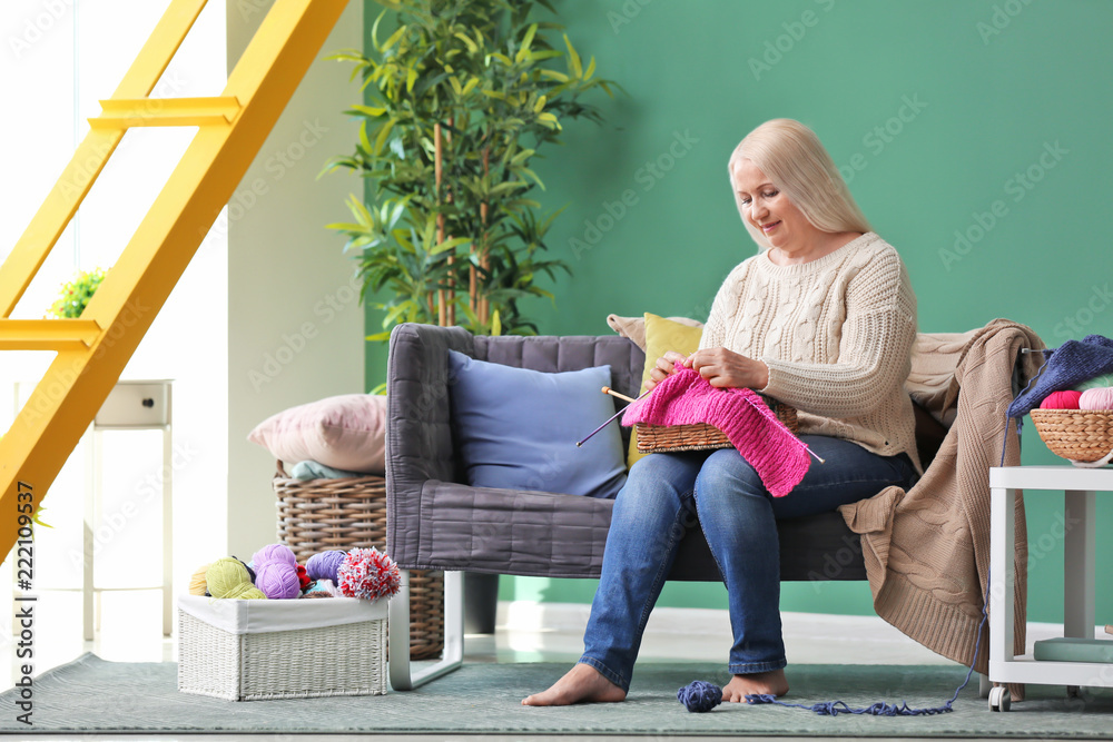 Mature woman sitting on sofa and knitting warm sweater indoors