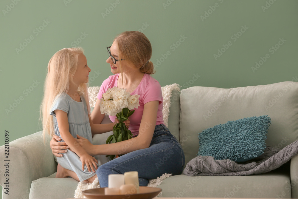 Cute little girl and her mother with beautiful flowers sitting on sofa at home