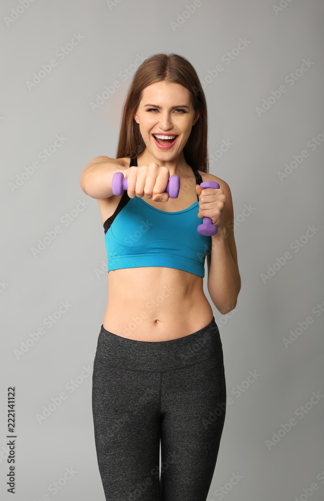 Sporty young woman with dumbbells on grey background