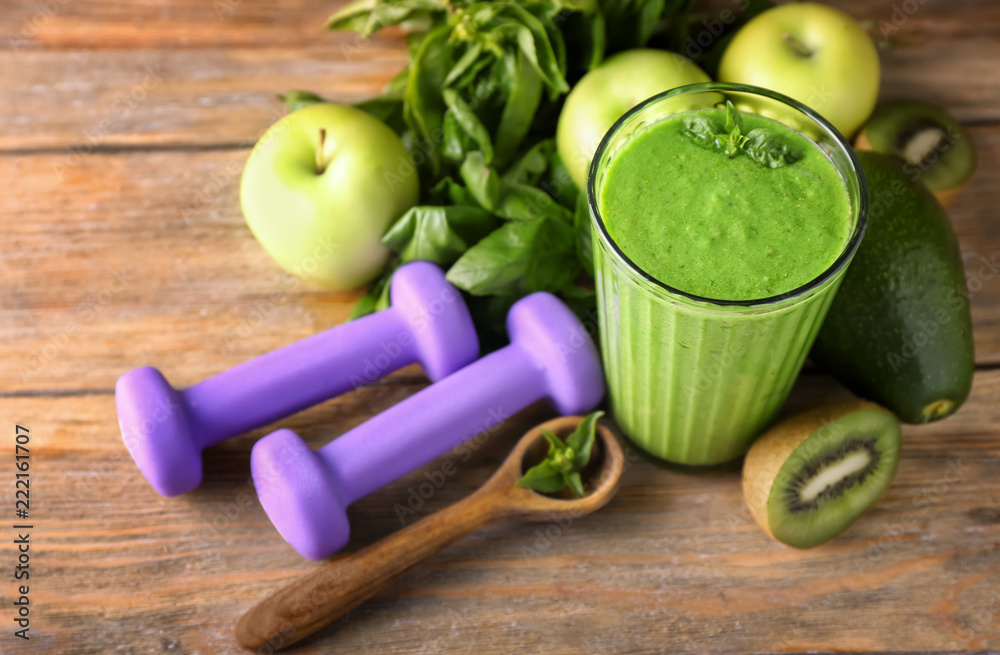 Glass of tasty green smoothie with ingredients and dumbbells on wooden table. Diet concept