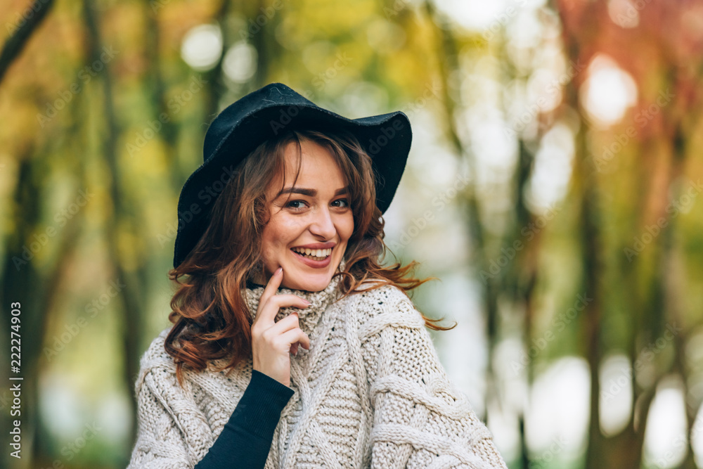 a girl in a black hat with a charming smile stands in the park on the background of autumn nature