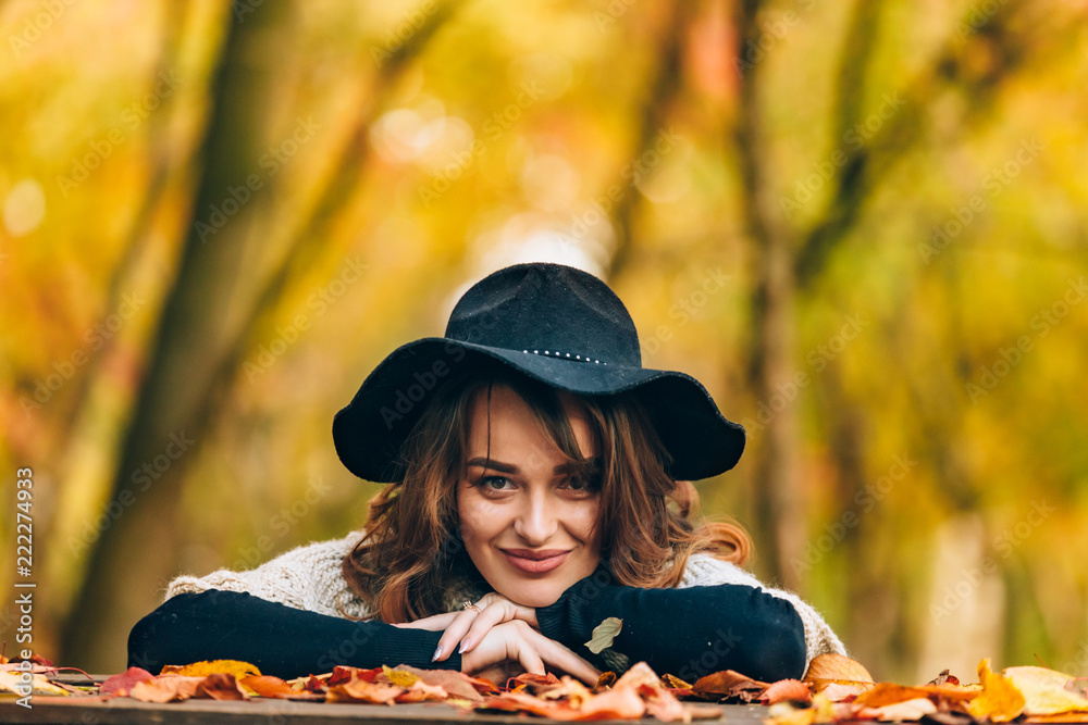 brown-haired woman in hat smiles and bases her hands on the table with foliage in the park