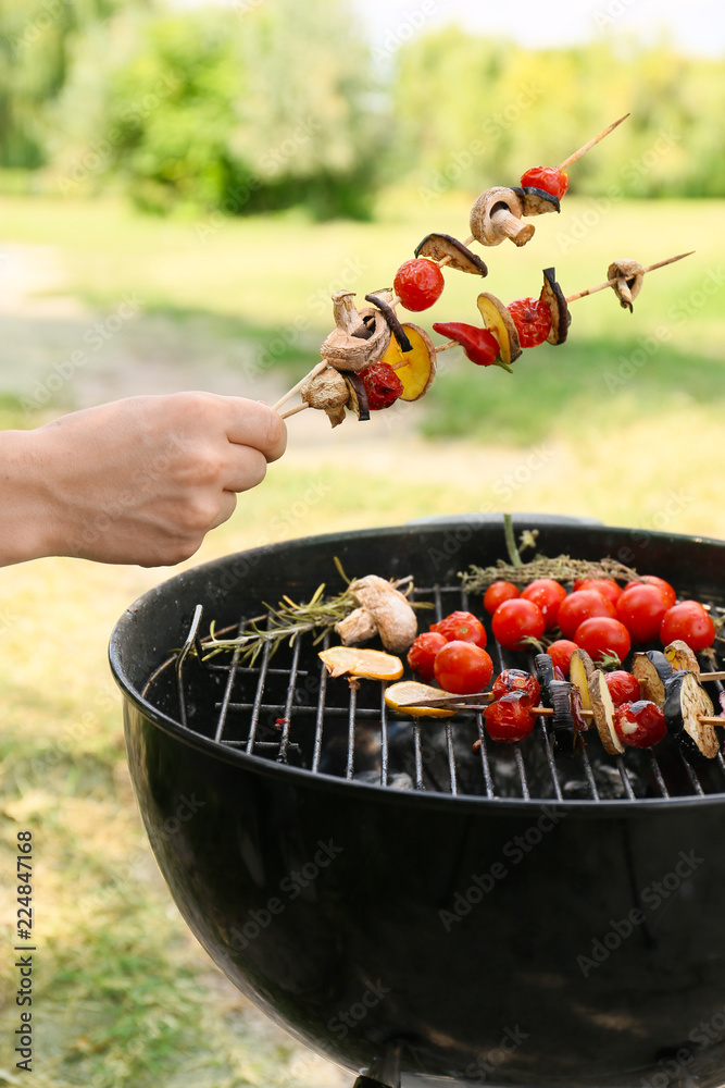 Man holding skewers with vegetables over barbecue grill outdoors