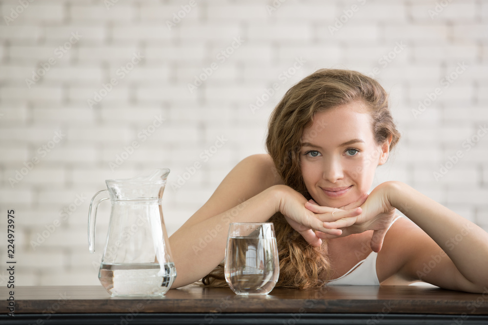 Beautiful young woman in joyful postures with jug and glass of drinking water on the side