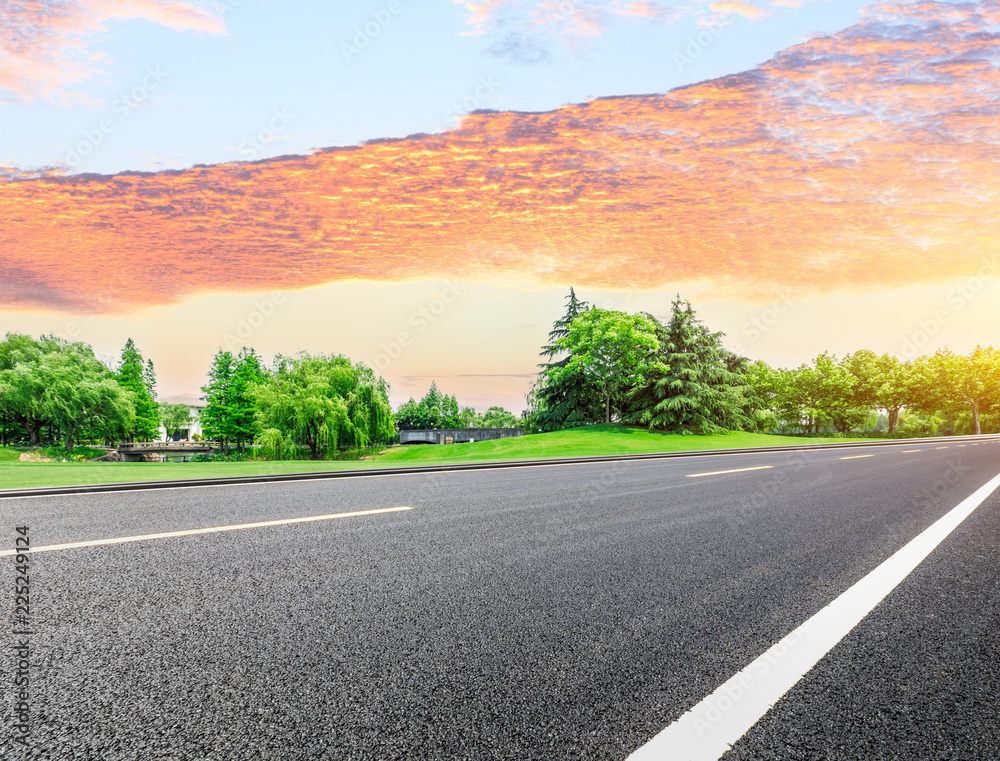 Empty asphalt road and green forest with colorful clouds at sunset