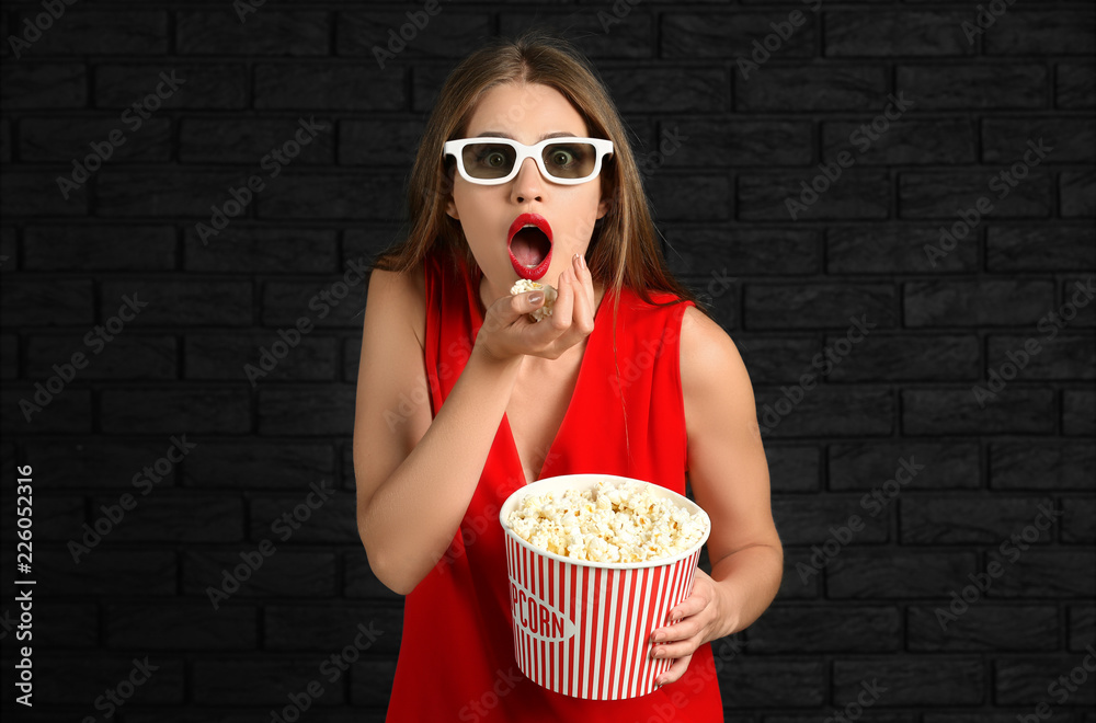 Emotional young woman with cup of popcorn and 3D cinema glasses against dark brick wall