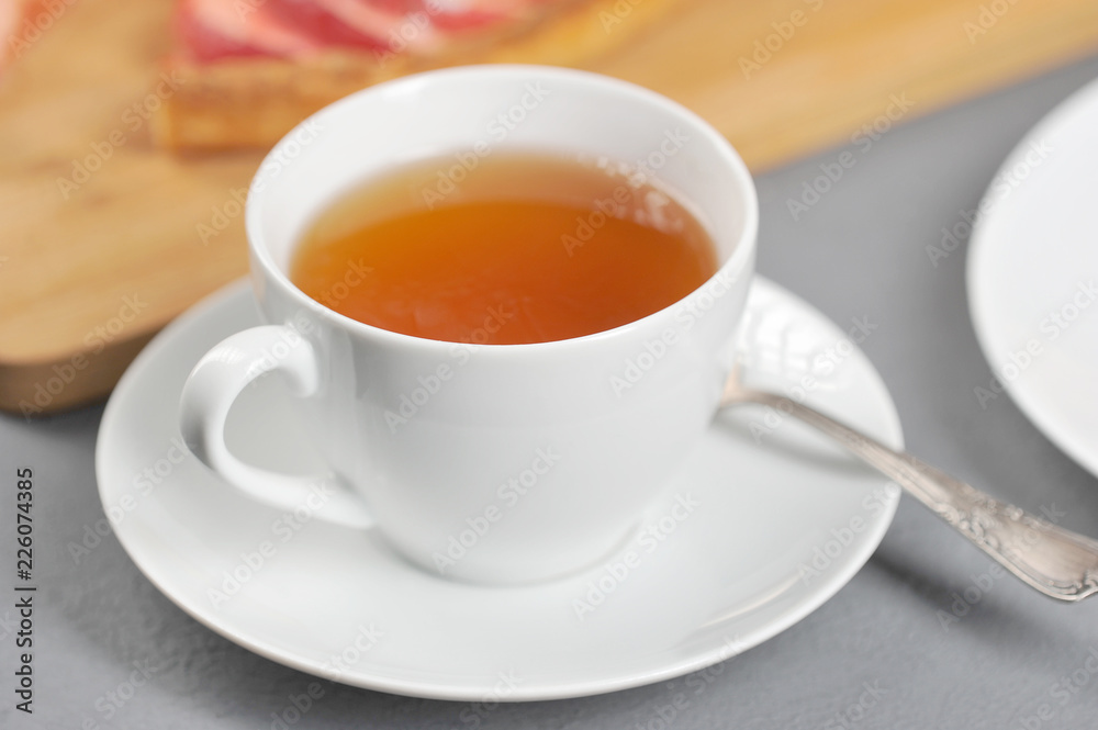 White cup with tea. On a saucer a teaspoon. In the background is an apple pie. Light background. Clo