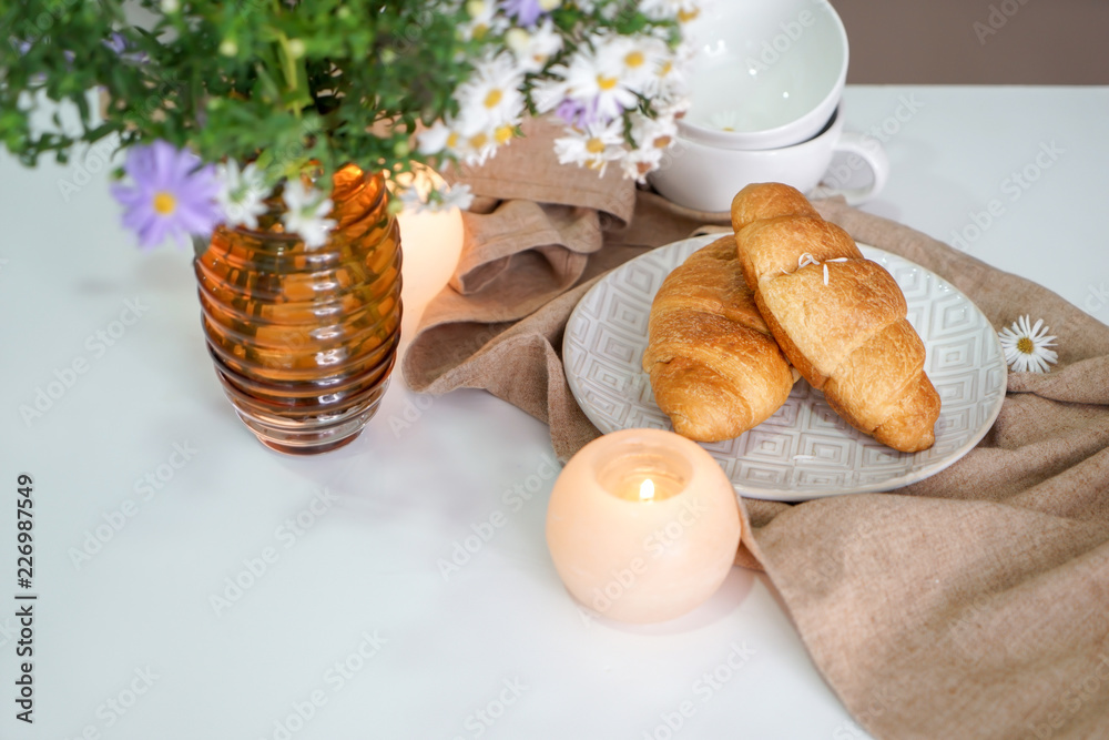 Burning candles with croissants and flowers on light table