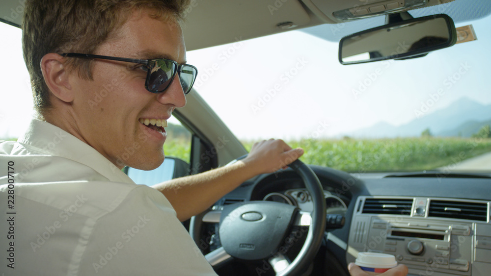 CLOSE UP: Smiling Caucasian man holds a takeaway cup of coffee while driving.