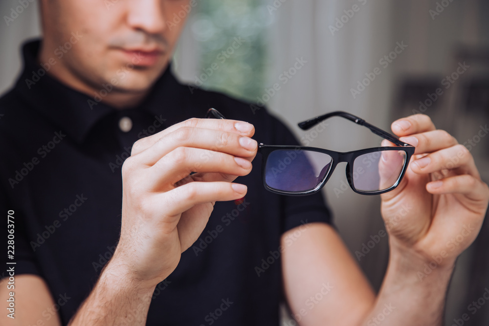 young man holds stylish black-rimmed eyeglasses and examines it in the room. Close-up