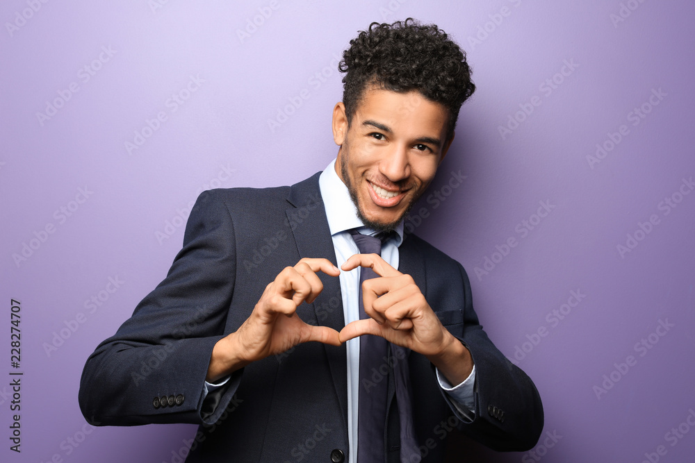 Handsome African-American businessman making heart with his hands on color background