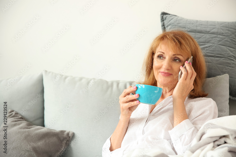 Beautiful mature woman drinking coffee while talking by phone at home